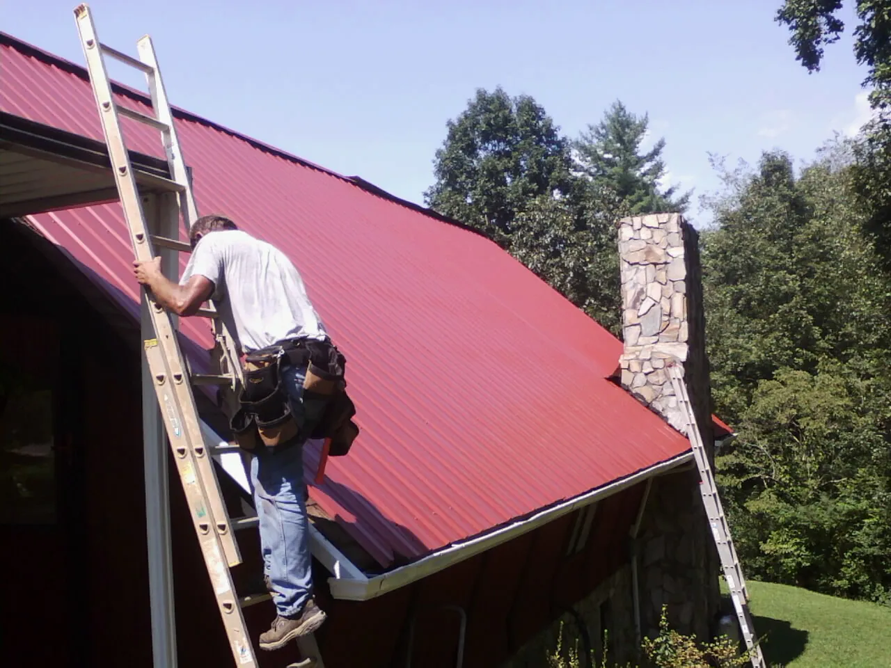 Roofer on a ladder beside a red metal roof and stone chimney — Soffit & Fascia Repair by Roofing Brothers in Kilmarnock